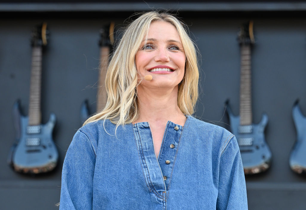 Cameron Diaz wears a casual denim outfit and smiles while standing on stage in front of a display of electric guitars