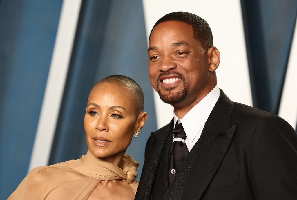Jada Pinkett Smith and Will Smith pose together at an event. Jada wears a sheer, high-neck gown, and Will wears a tuxedo