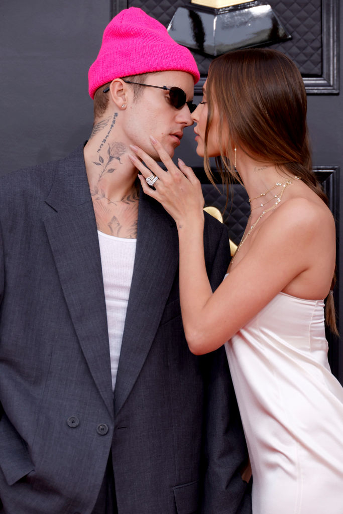 Justin Bieber in oversized suit, tank top, beanie, and sunglasses. Hailey Bieber in satin dress, holding his face, noses touching