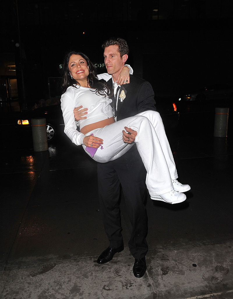 A man in a suit carries Bethenny Frankel, who is wearing a white outfit, in his arms on a street at night