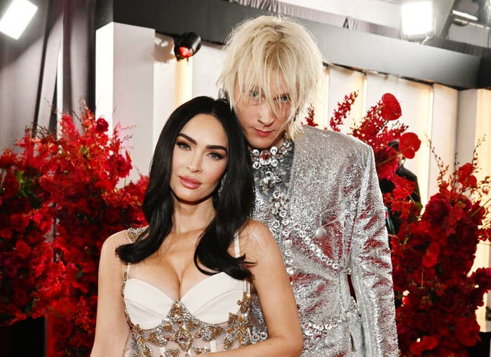 Megan Fox and Machine Gun Kelly on the red carpet. Megan is wearing a white gown with silver embellishments, and Machine Gun Kelly is in a silver metallic suit