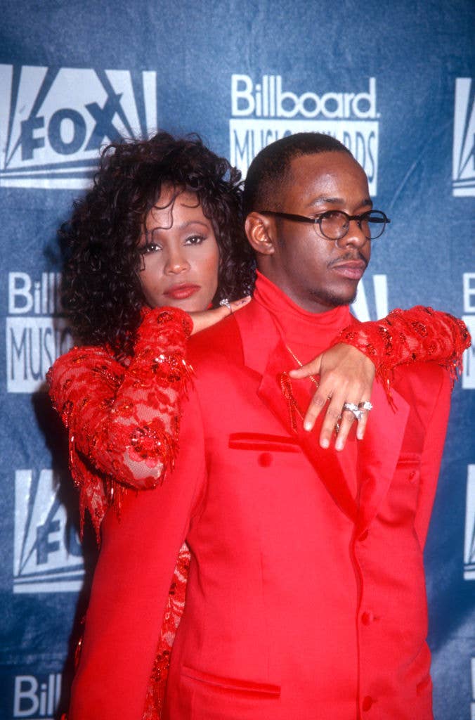 Whitney Houston and Bobby Brown at the Billboard Music Awards. Whitney wears a sequined dress, and Bobby wears a red suit and glasses