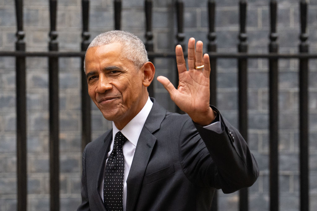 Barack Obama waves with a smile, dressed in a black suit, white dress shirt, and polka dot tie, standing in front of a wrought iron fence