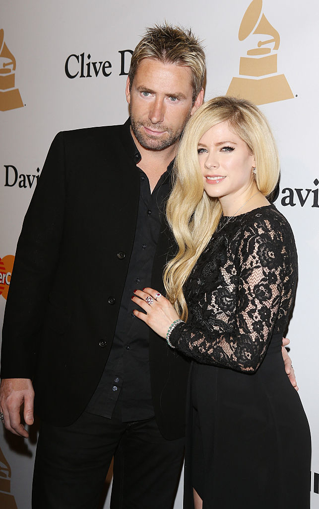 Chad Kroeger and Avril Lavigne, with Chad wearing a black suit jacket and shirt, and Avril in a black lace dress, posing together at an event with Grammy logos