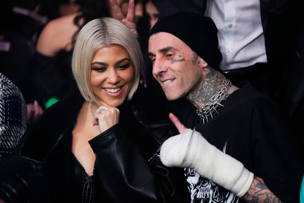 Kourtney Kardashian and Travis Barker smile together at an event. Travis shows a thumbs-up with an arm in a white cast