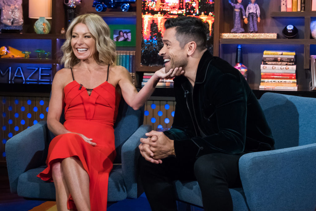 Kelly Ripa and Mark Consuelos are seated on a talk show set. Kelly Ripa is in a sleeveless dress and Mark Consuelos is in a dark jacket