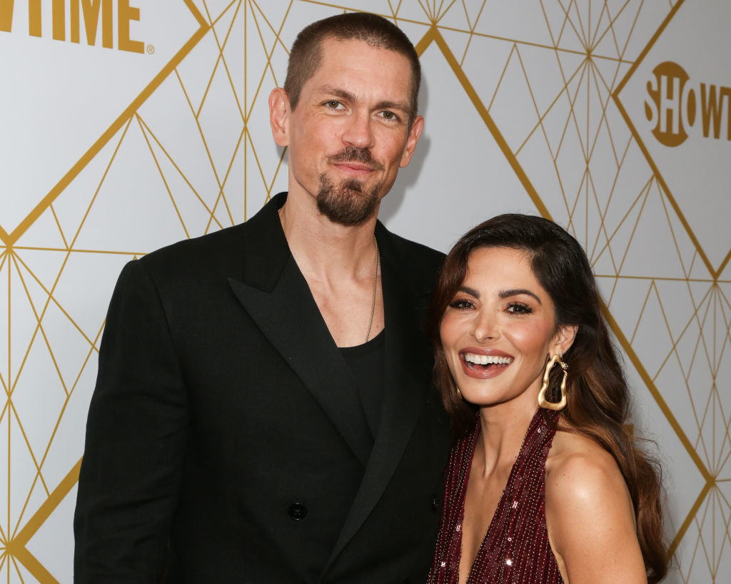 A man in a black suit and a woman in a sleeveless, sparkling dress stand smiling on a red carpet background with the Showtime logo