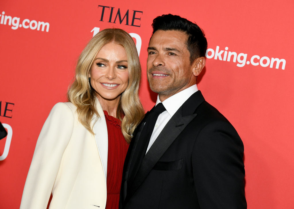 Kelly Ripa and Mark Consuelos pose together on the red carpet at an event. Kelly wears a chic dress with a white blazer; Mark is in a classic black suit and tie