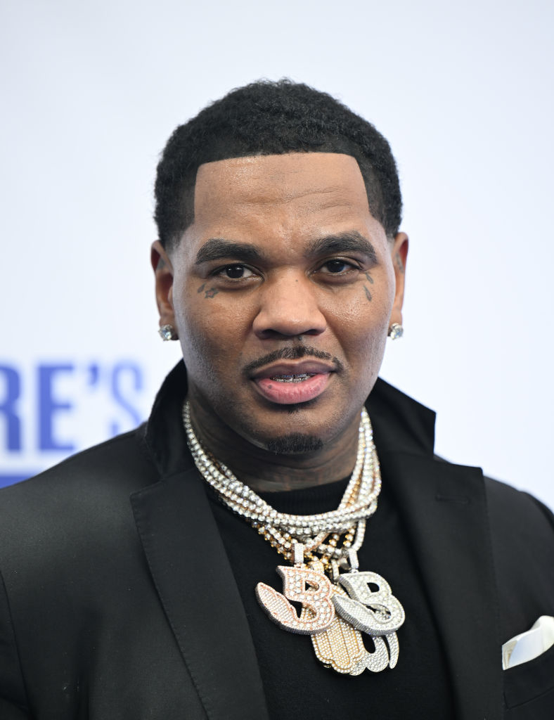 Kevin Gates wearing a black jacket and white shirt, adorned with multiple diamond necklaces and earrings, attends an event