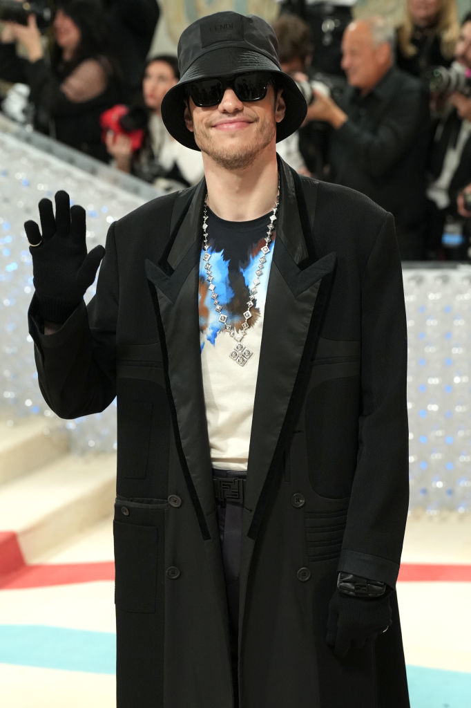 Pete Davidson on a red carpet wearing a black bucket hat, dark sunglasses, a long black coat, black gloves, a graphic t-shirt, and a silver chain necklace