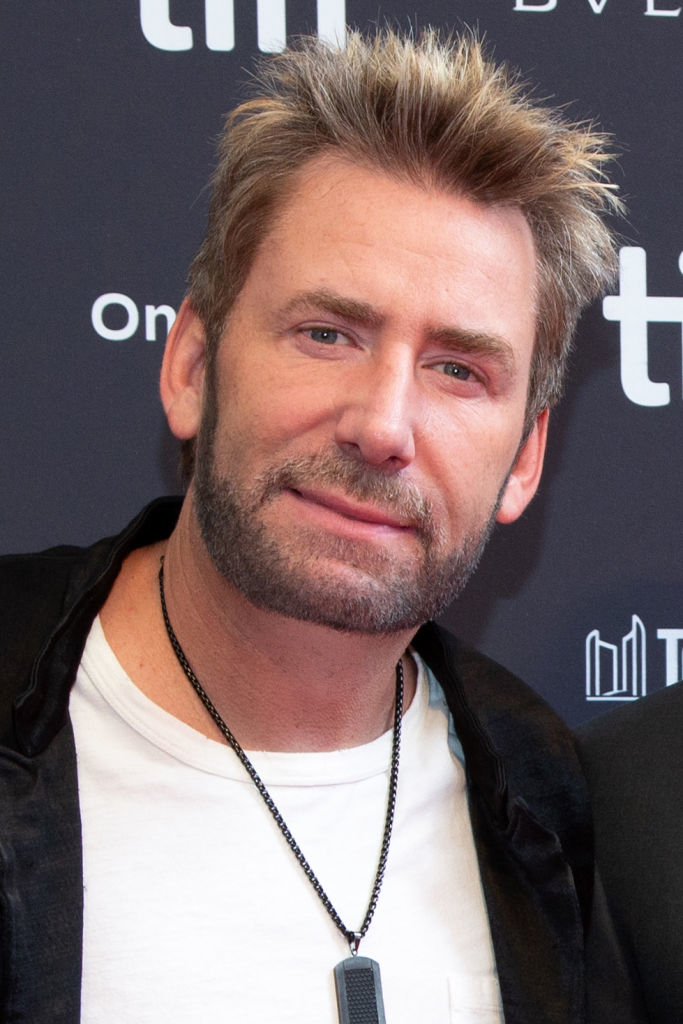 A man with spiked hair and a beard, wearing a white shirt with a necklace and a black jacket, poses at a media event