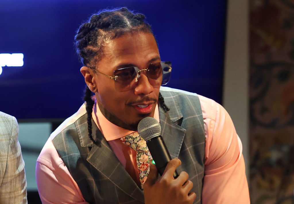 Nick Cannon holding a microphone, wearing a pink shirt with a floral tie and a grey checkered vest