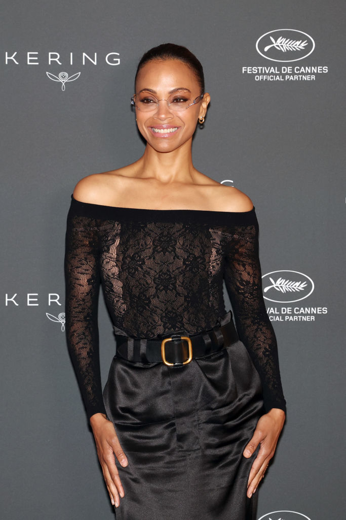 Zoe Saldana on the red carpet wearing an off-shoulder lace top and high-waisted belted trousers, smiling. Background has Kering and Cannes festival logos