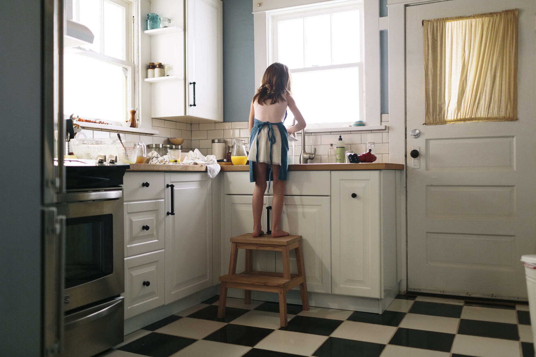 A child stands on a wooden stool at a kitchen counter, reaching towards a window. The kitchen has white cabinets and a checkered floor