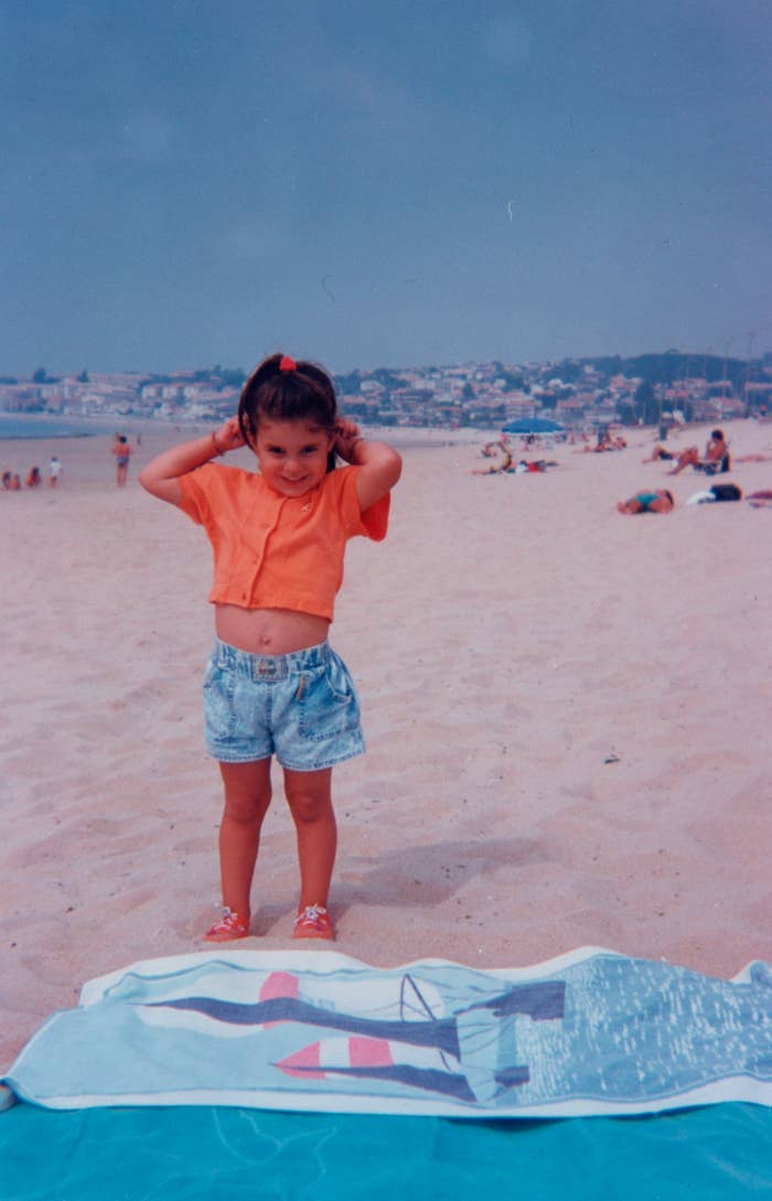 A young girl stands on a sandy beach wearing a short-sleeve shirt, denim shorts, and sandals. She is posing in front of a beach towel, with people and buildings in the background