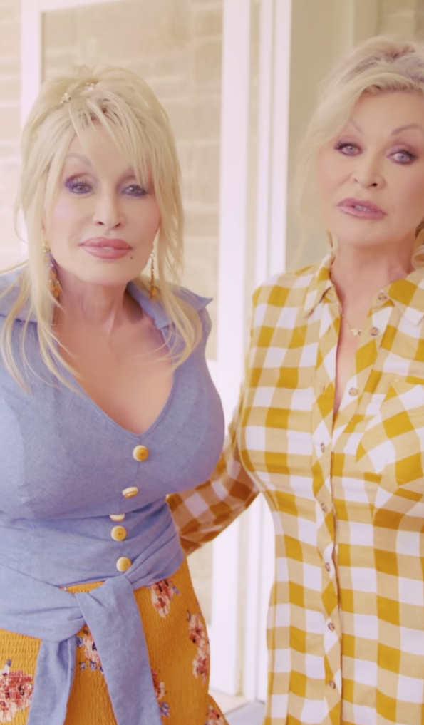 Dolly Parton and another woman stand together, smiling. Dolly is wearing a blue top and floral skirt. The other woman wears a checked shirt