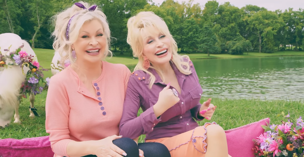 Dolly Parton and another woman sitting on a blanket by a lake, smiling and laughing, with flowers and a llama in the background