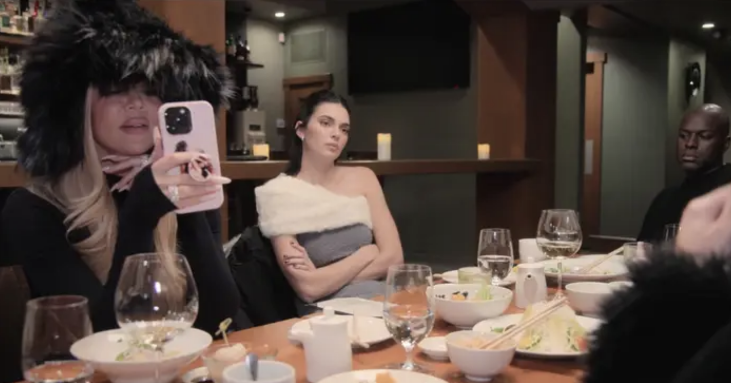 Khloé Kardashian, Kendall Jenner, and another person sit at a dining table with various dishes, Khloé taking a photo with her phone