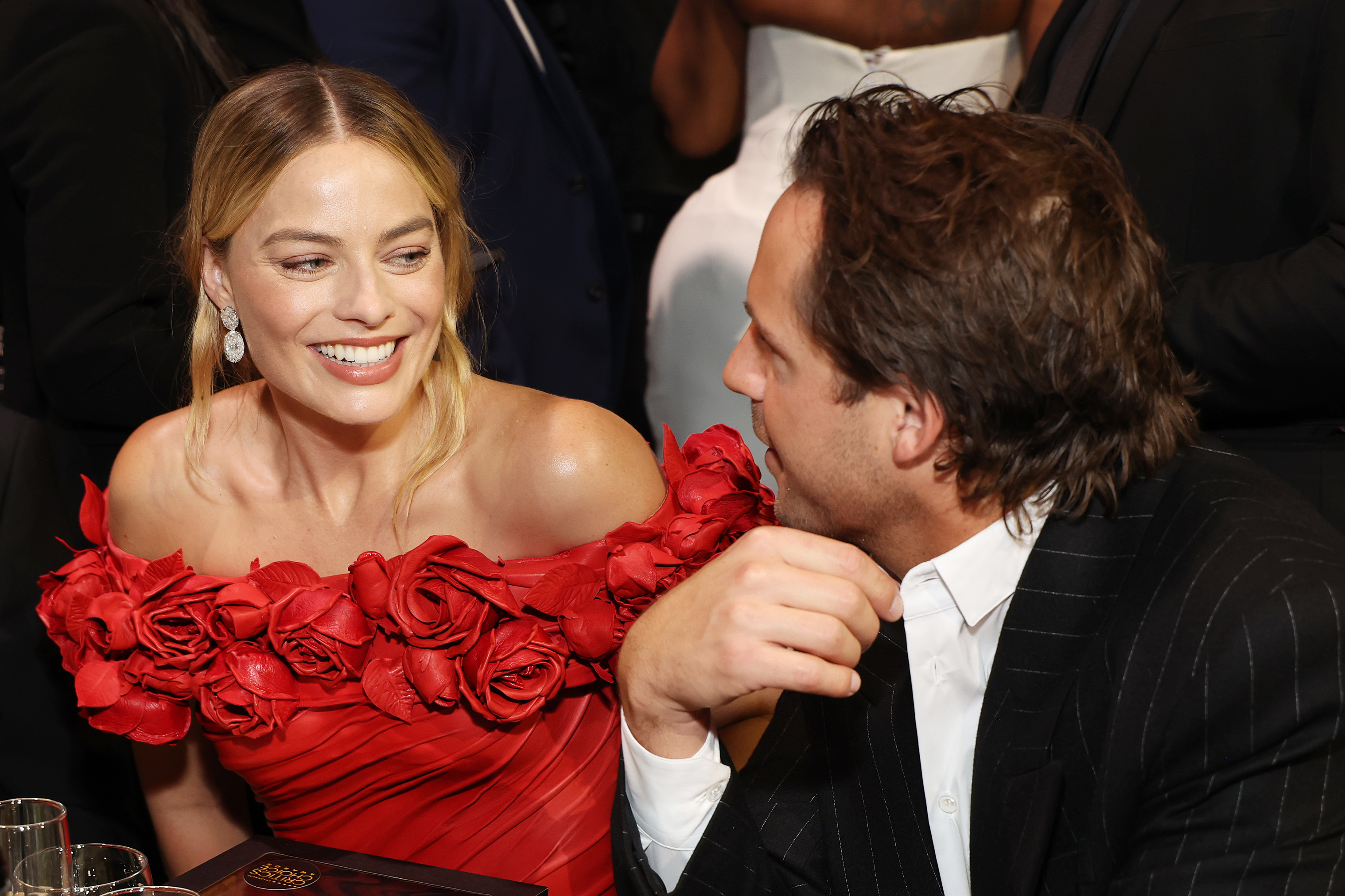 Margot Robbie in a floral off-the-shoulder dress smiles at a man in a striped suit at an event