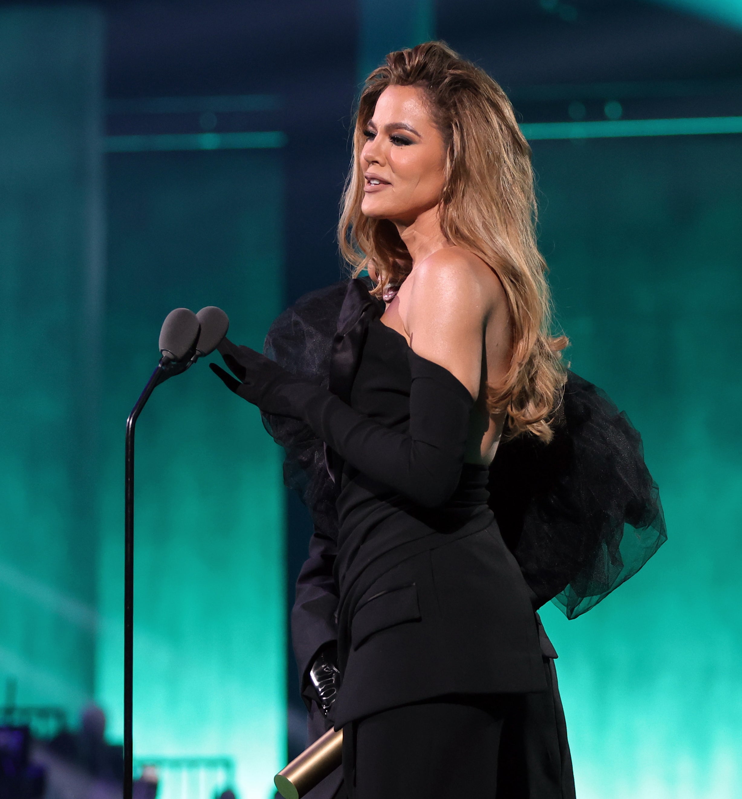 Khloé Kardashian stands on stage holding a microphone, wearing an elegant one-sleeve gown with volumized shoulder detail
