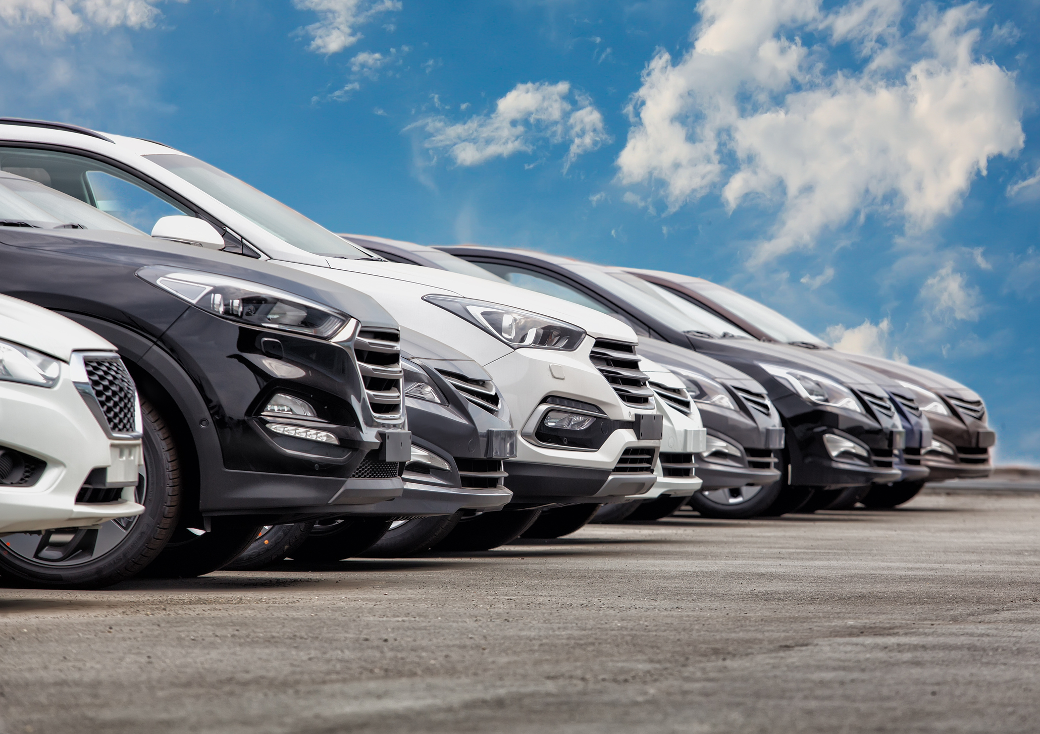 Several parked SUVs are aligned in a row on a concrete surface, with a clear blue sky and clouds visible in the background