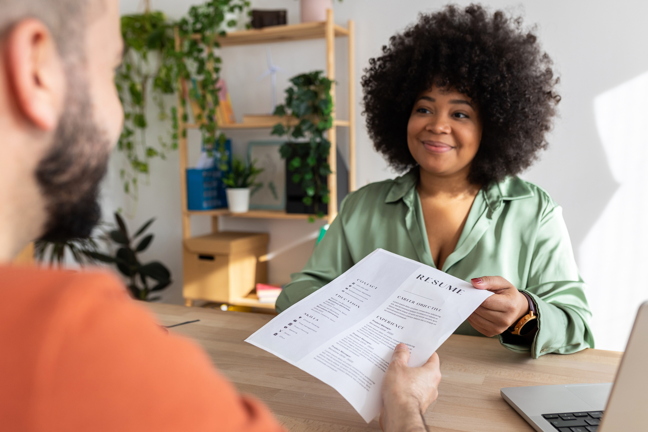 A person in a green blouse hands a resume to another person across a table in what appears to be an office setting. The woman is smiling