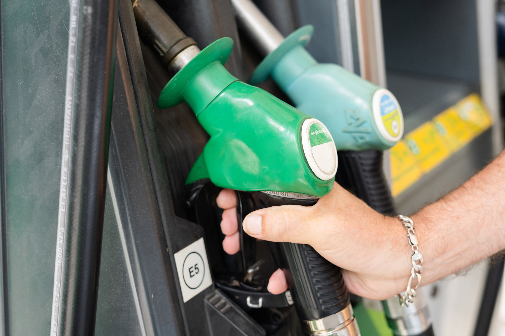 A hand holds a green fuel pump nozzle next to another nozzle at a gas station