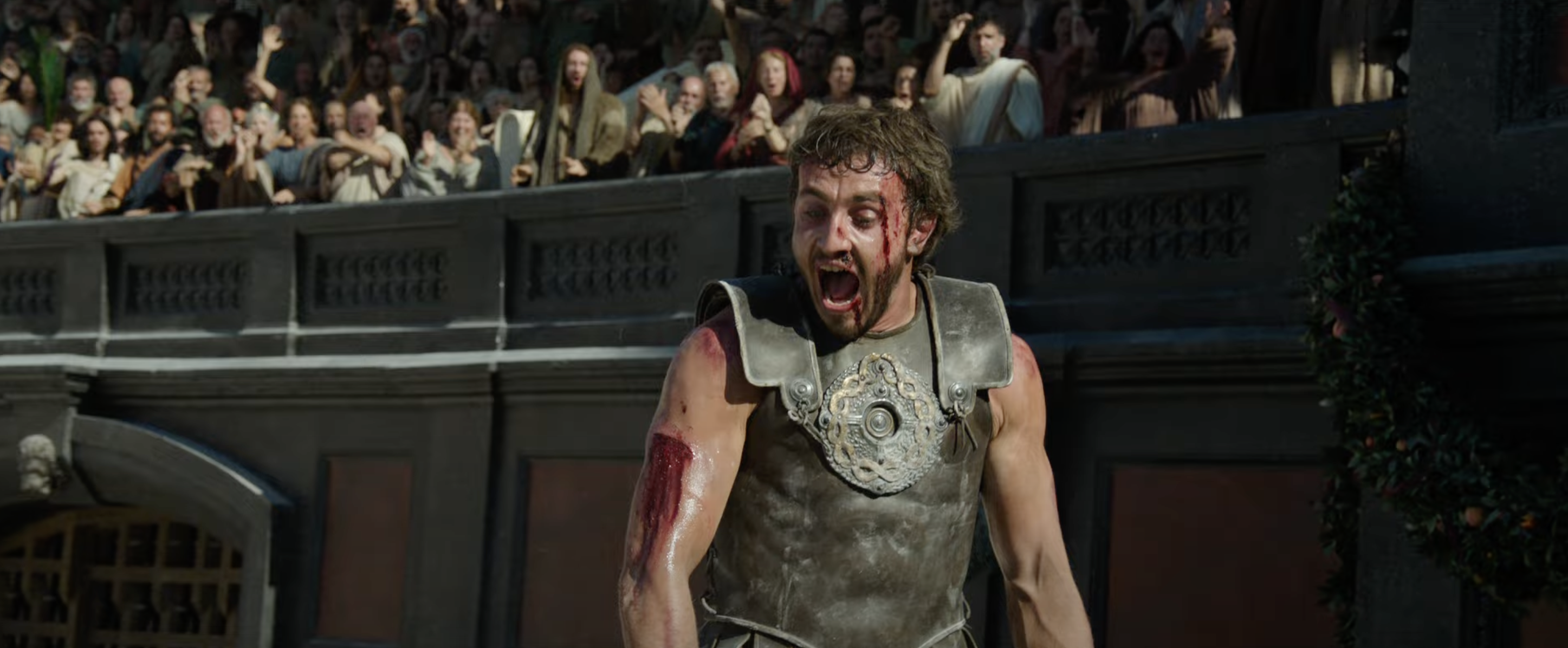 Paul Mescal, in gladiator armor with a bloody face, looks exhausted while standing in an arena. An audience in the background appears to be cheering