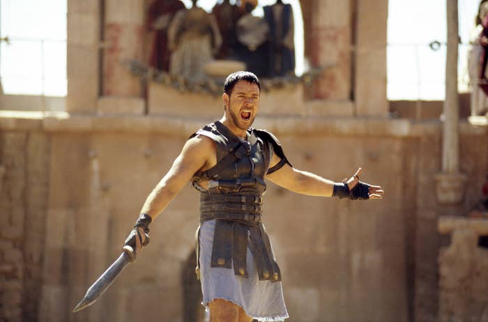 Russell Crowe, dressed as a gladiator in armor, stands in an ancient arena with arms outstretched and a sword in hand