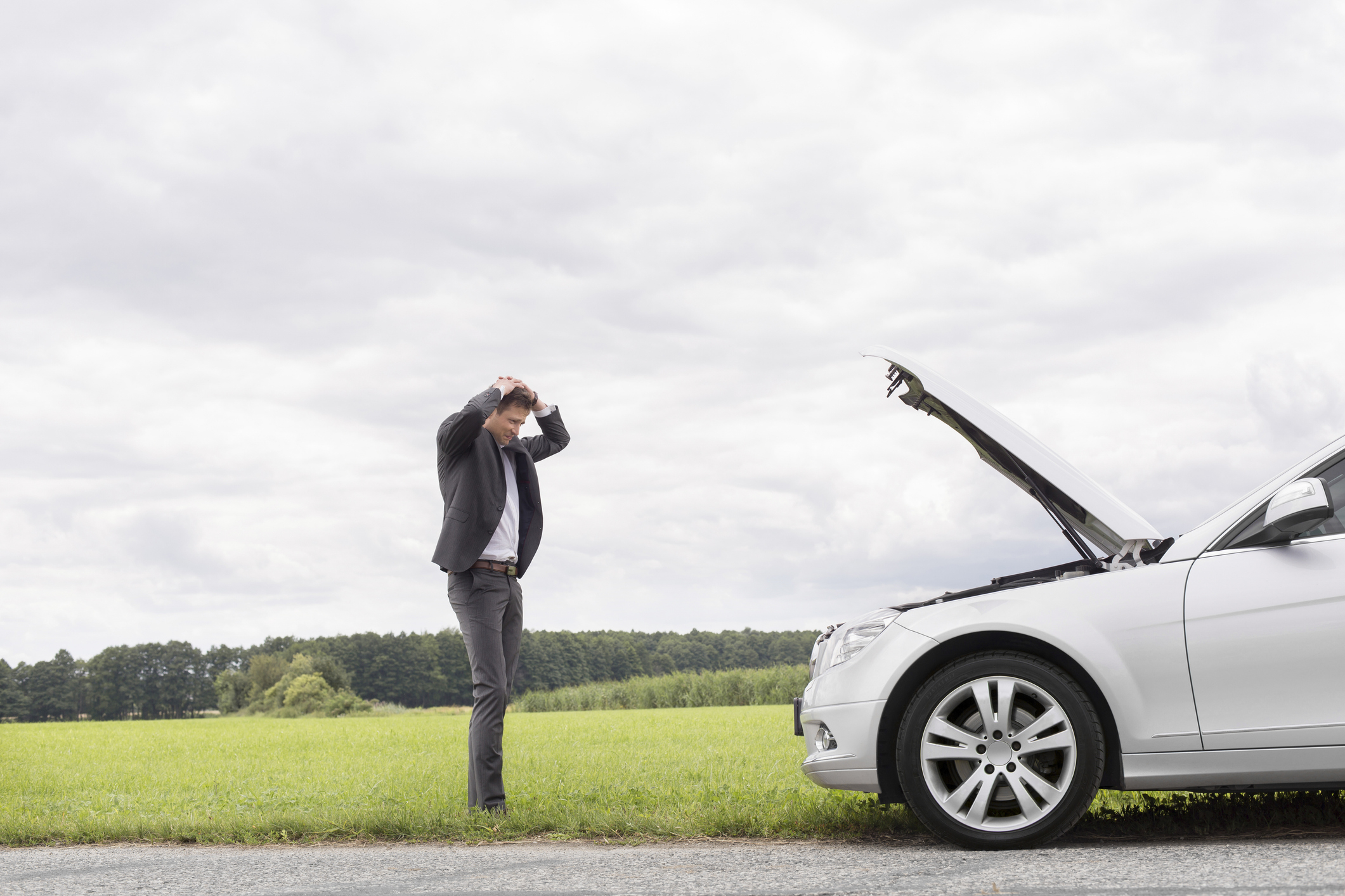A man in a suit stands on the side of the road with his hands on his head, looking at the open hood of a broken-down car in a rural area