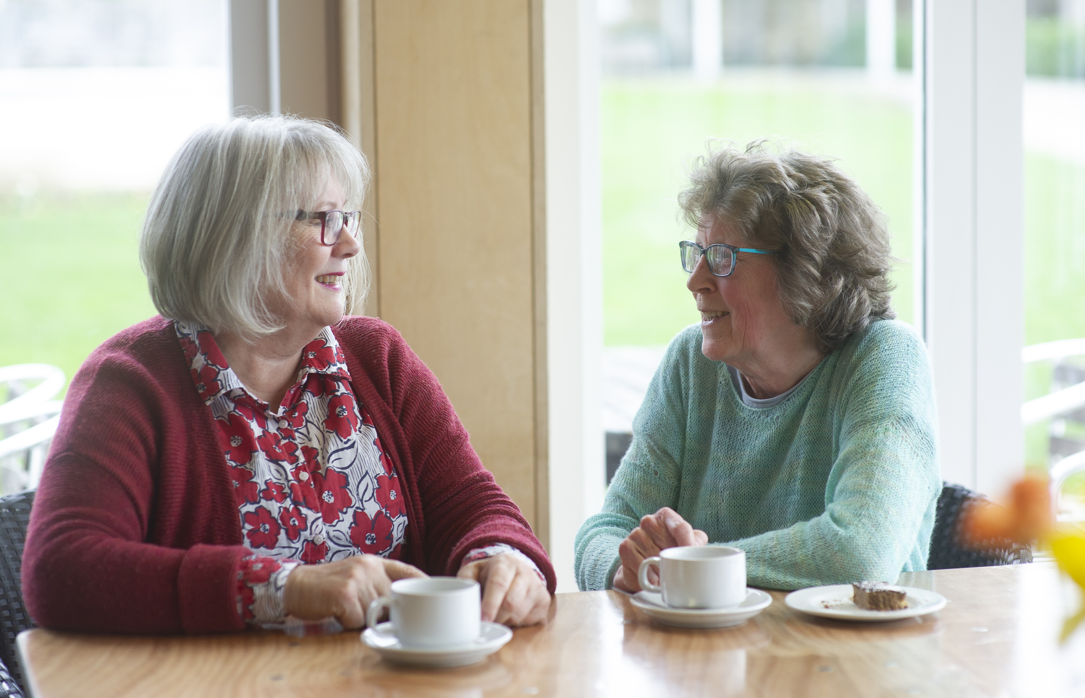 Two elderly women with short gray hair chatting over coffee at a table by a window. One wears a red cardigan and floral shirt, the other a turquoise sweater