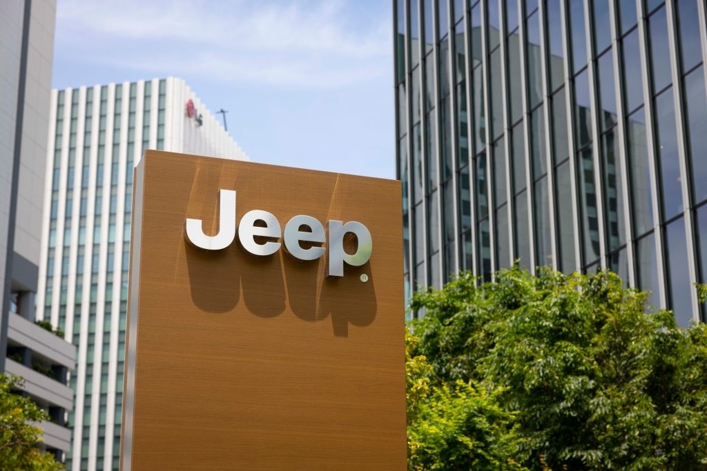 A Jeep sign stands amid a city setting with tall buildings and greenery in the background