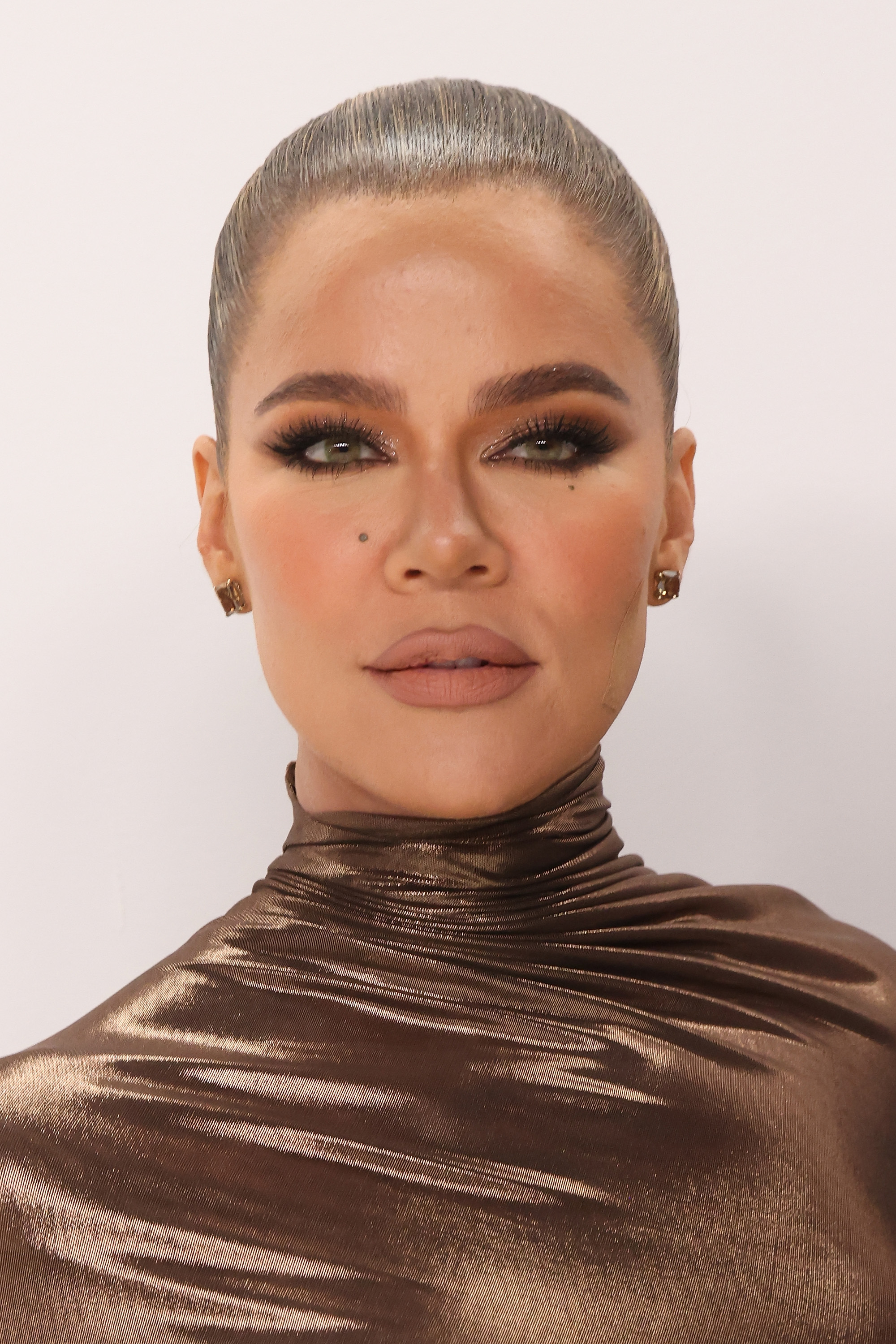 Khloé Kardashian in a metallic, high-neck dress, with her hair slicked back and wearing bold makeup