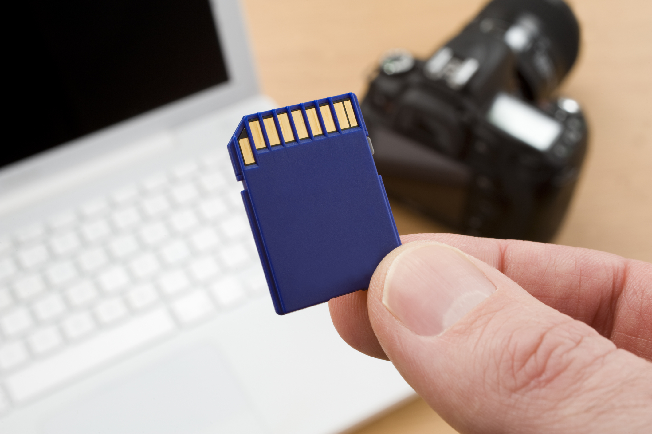 A close-up of a hand holding an SD card in front of a partially visible laptop and a blurred camera in the background