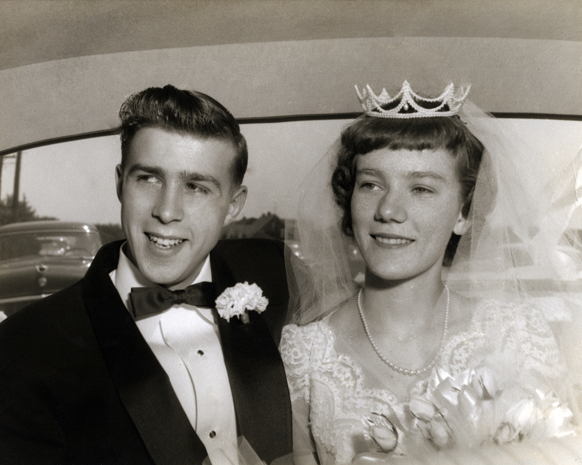 A young couple, man in a black tuxedo with a bow tie and boutonniere, woman in a wedding dress with a delicate tiara, sitting together in a car on their wedding day