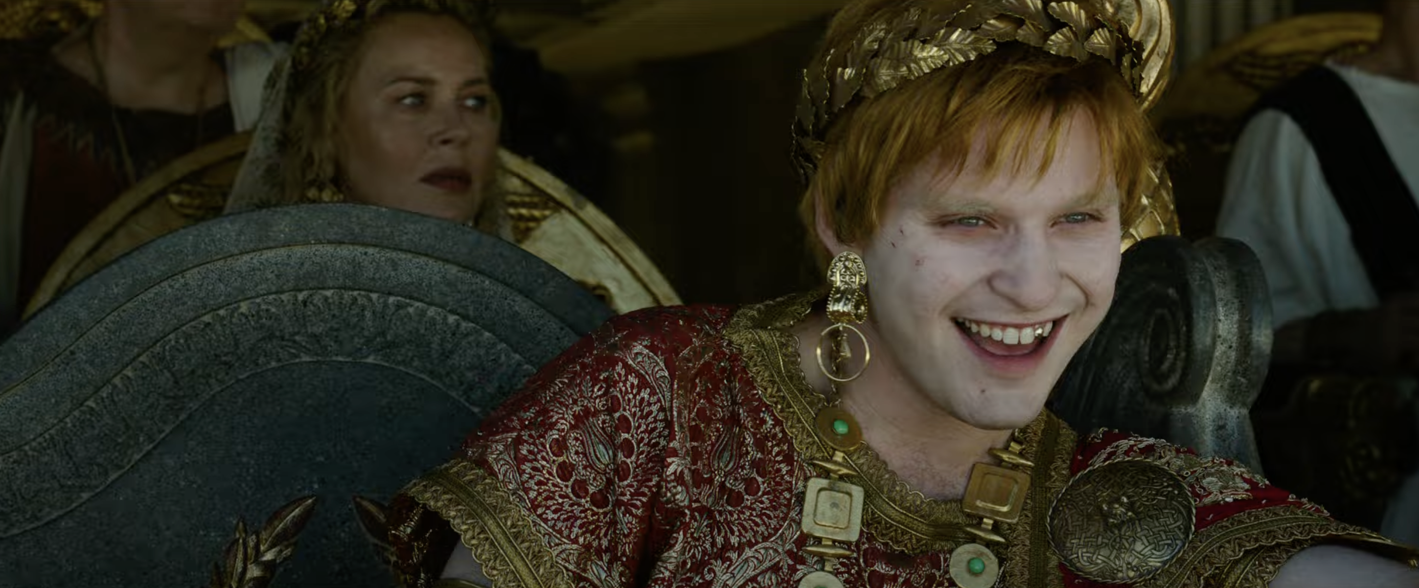 A person wears a Roman-style costume, including a decorative gold crown and ornate red attire, smiling while sitting on a throne. Another person is in the background