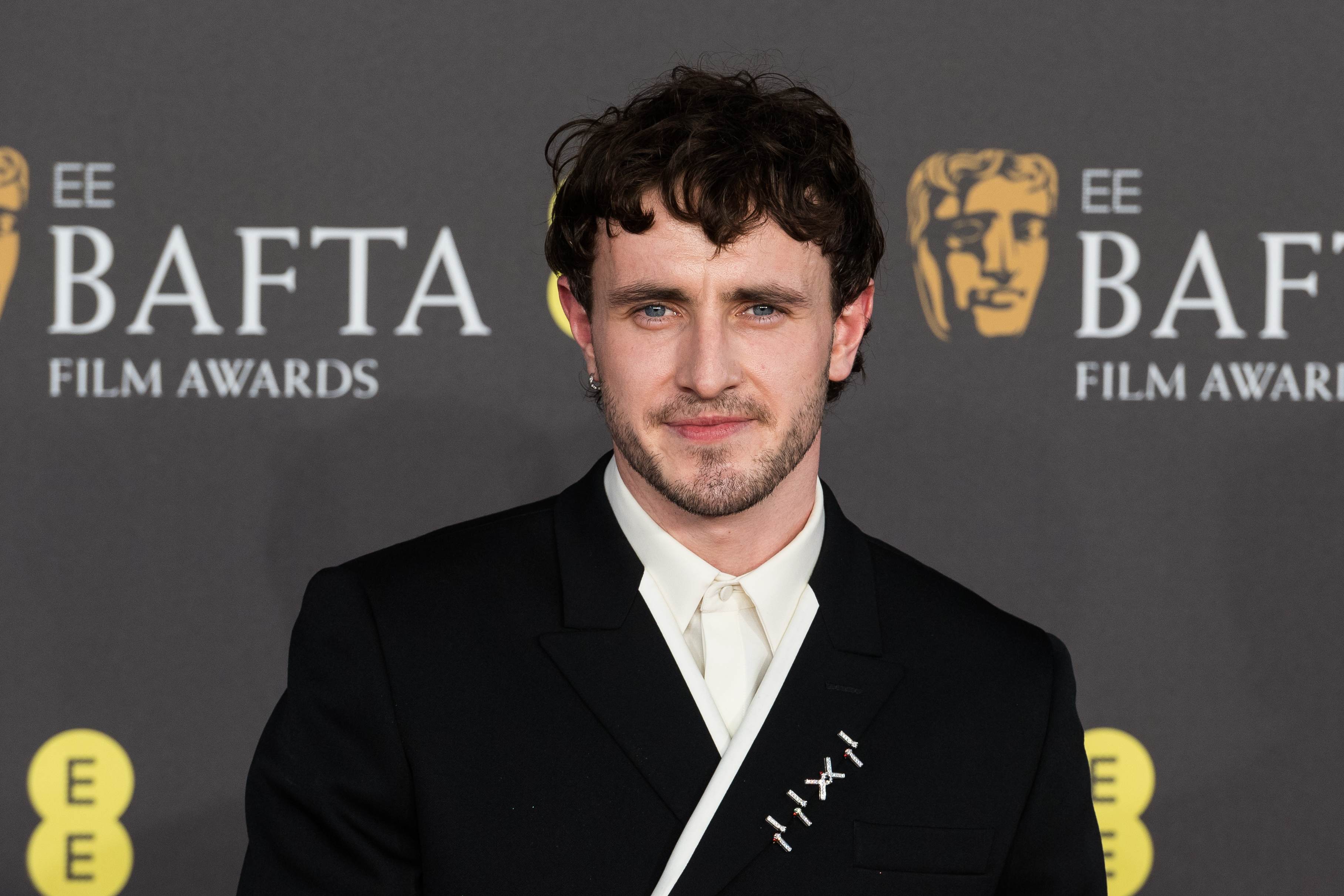 Paul Mescal at the BAFTA Film Awards, wearing a black suit with a white shirt. There is a BAFTA logo backdrop behind him