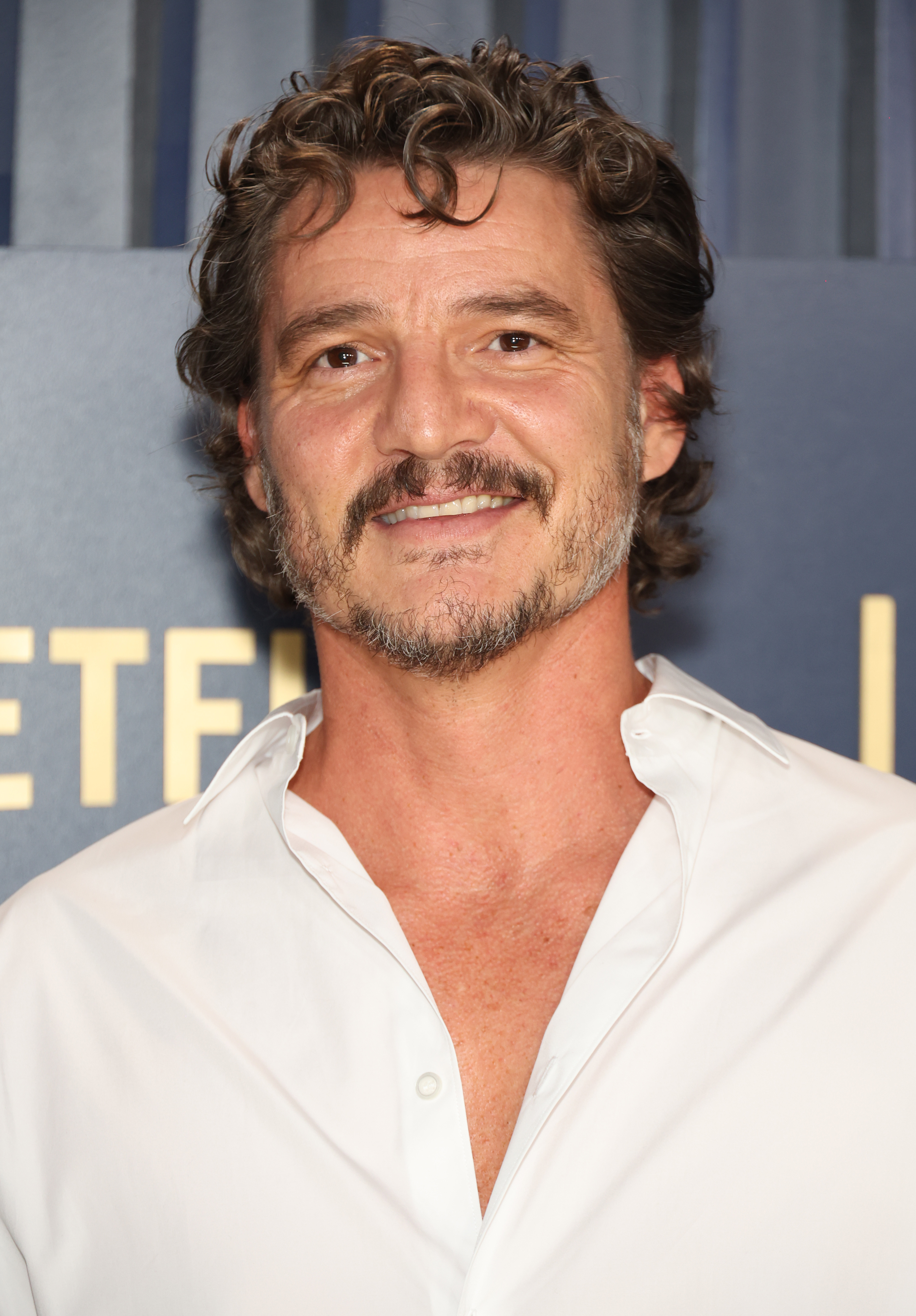 Pedro Pascal smiles, wearing a white shirt with an open collar, at a media event