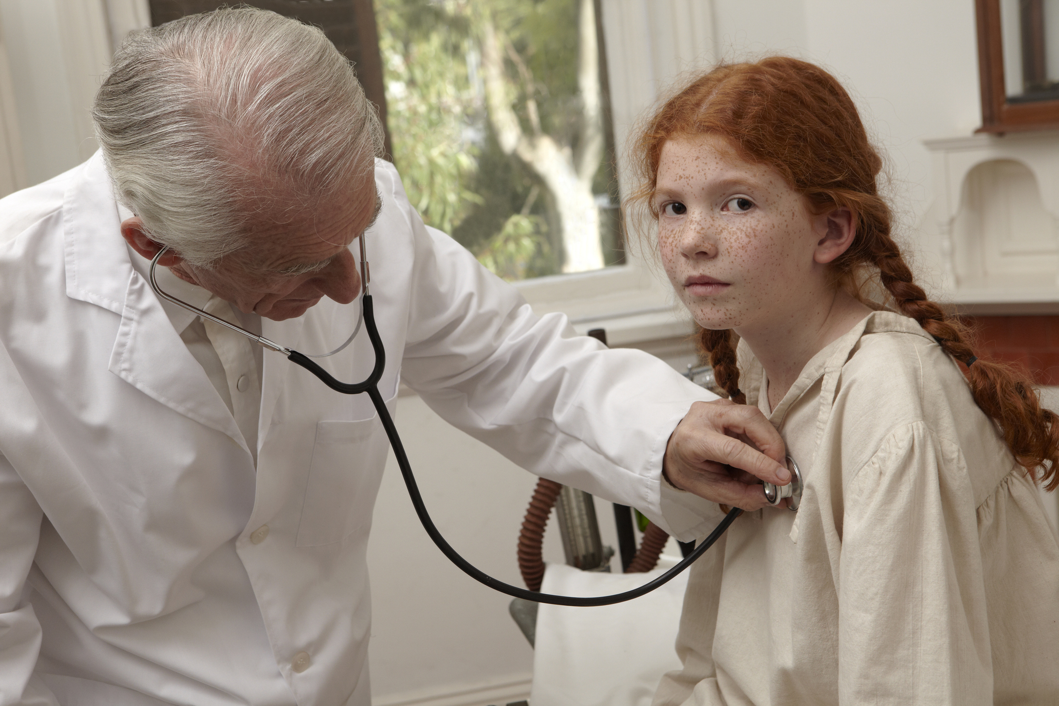 A doctor in a white coat uses a stethoscope to examine a girl with red hair and braids wearing a light shirt