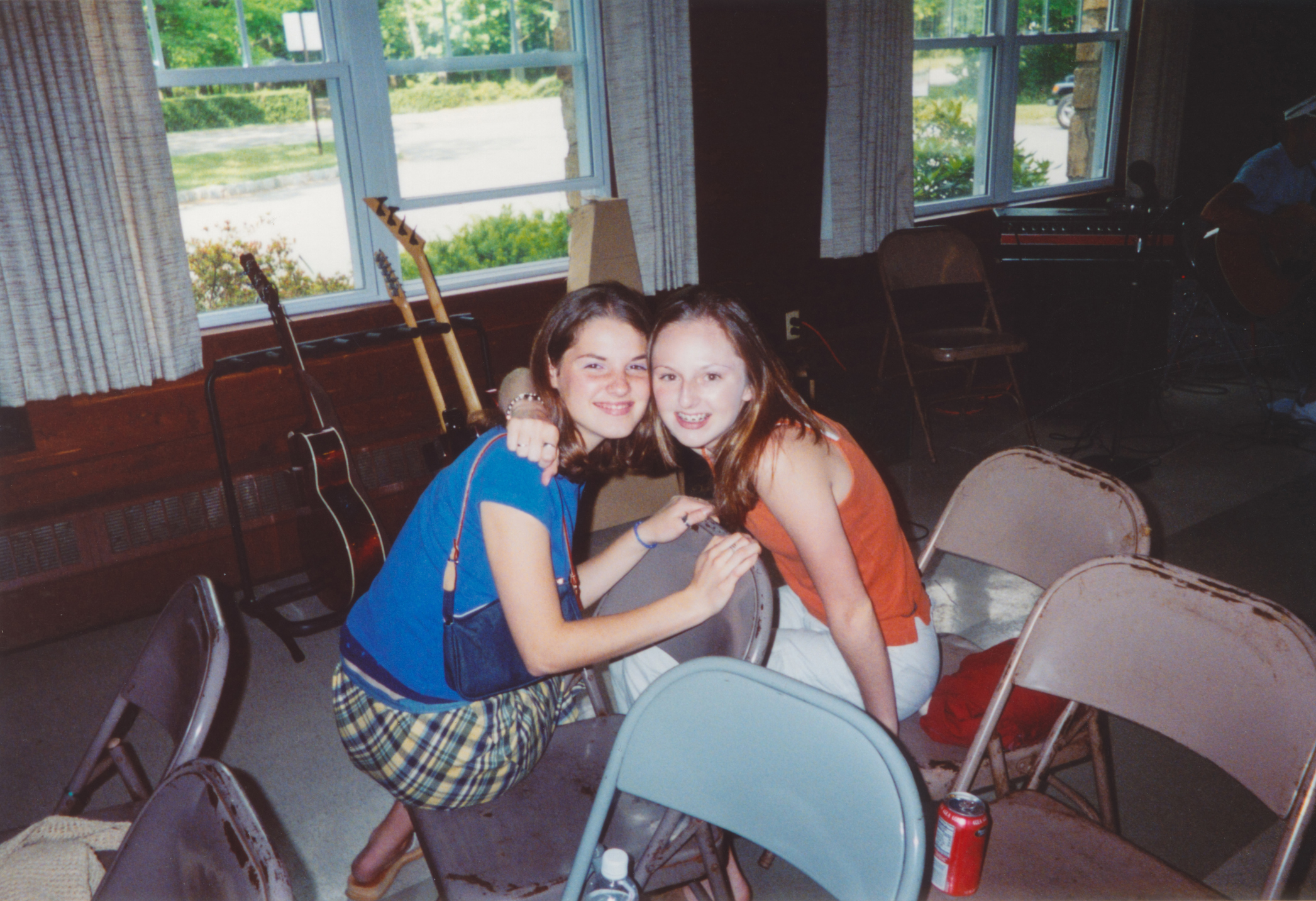 Two young women, one in a blue top and plaid skirt, and the other in an orange top and white pants, smile and pose for a photo in a music room with chairs and guitars