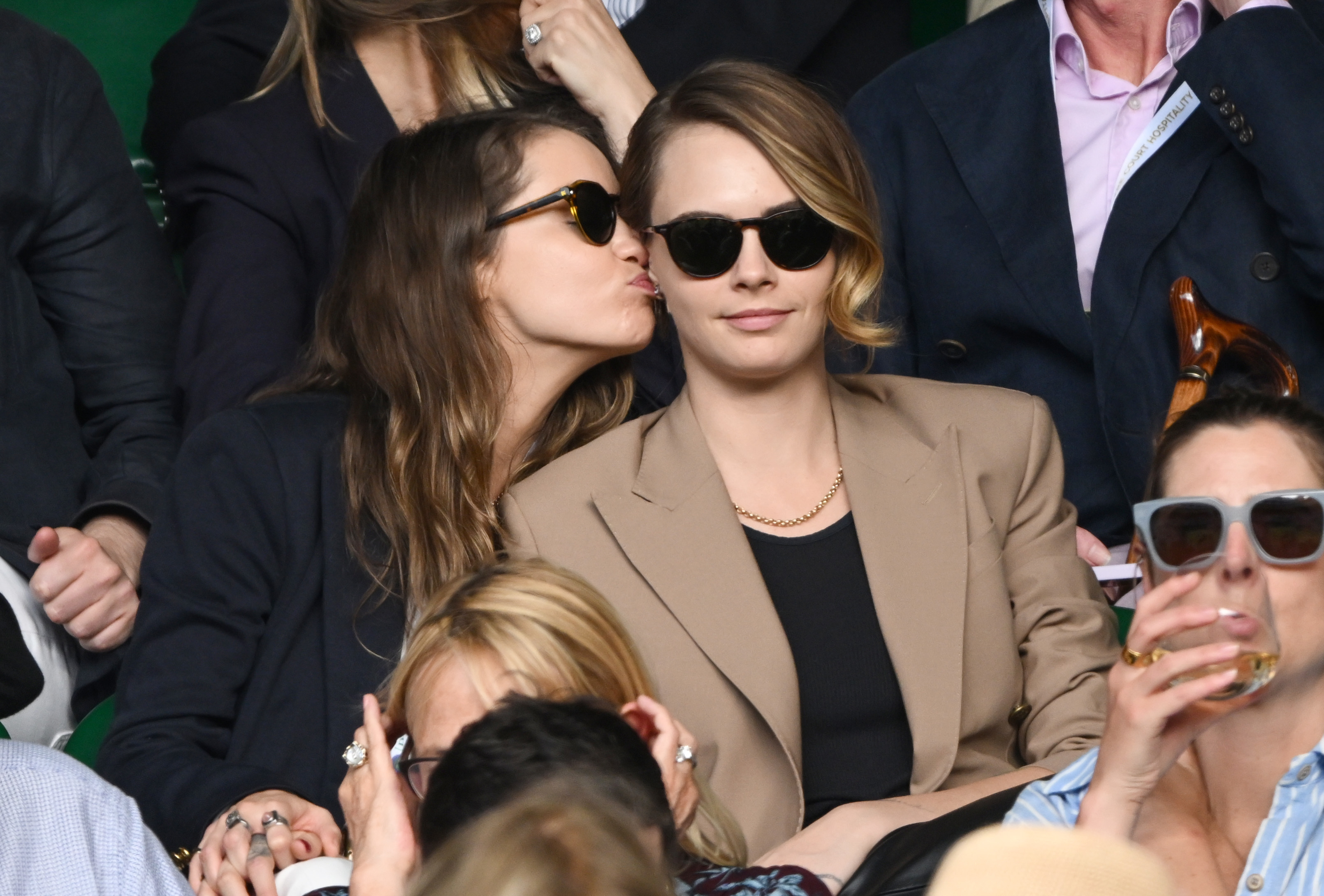 Minke kisses Cara Delevingne on the cheek while they sit at an event. Both wear sunglasses and stylish blazers. Other attendees are in the background