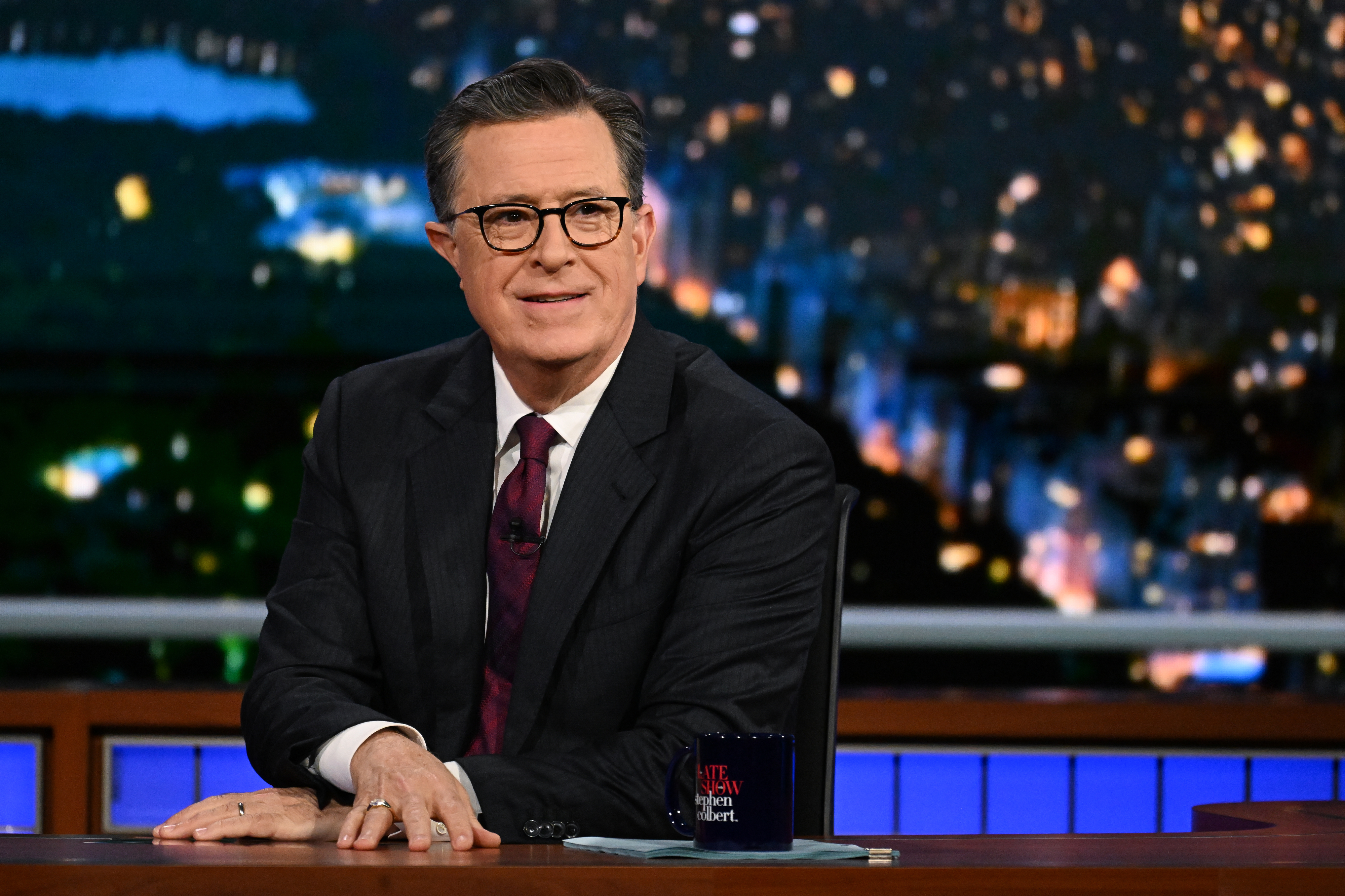 Stephen Colbert, in a dark suit and tie, sits at a desk with a cityscape background on &quot;The Late Show with Stephen Colbert.&quot;