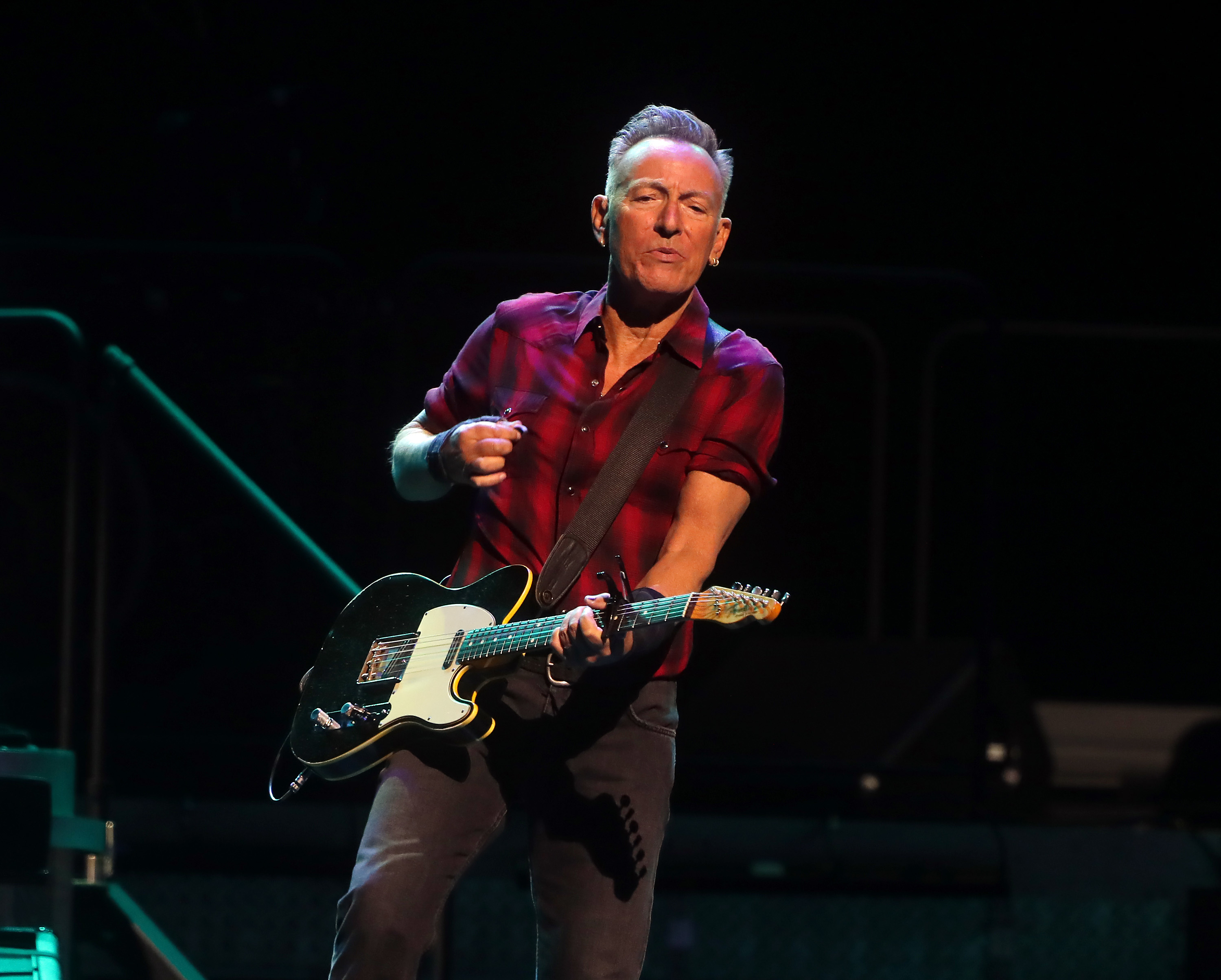 Bruce Springsteen plays an electric guitar on stage, wearing a short-sleeve, button-up shirt and jeans