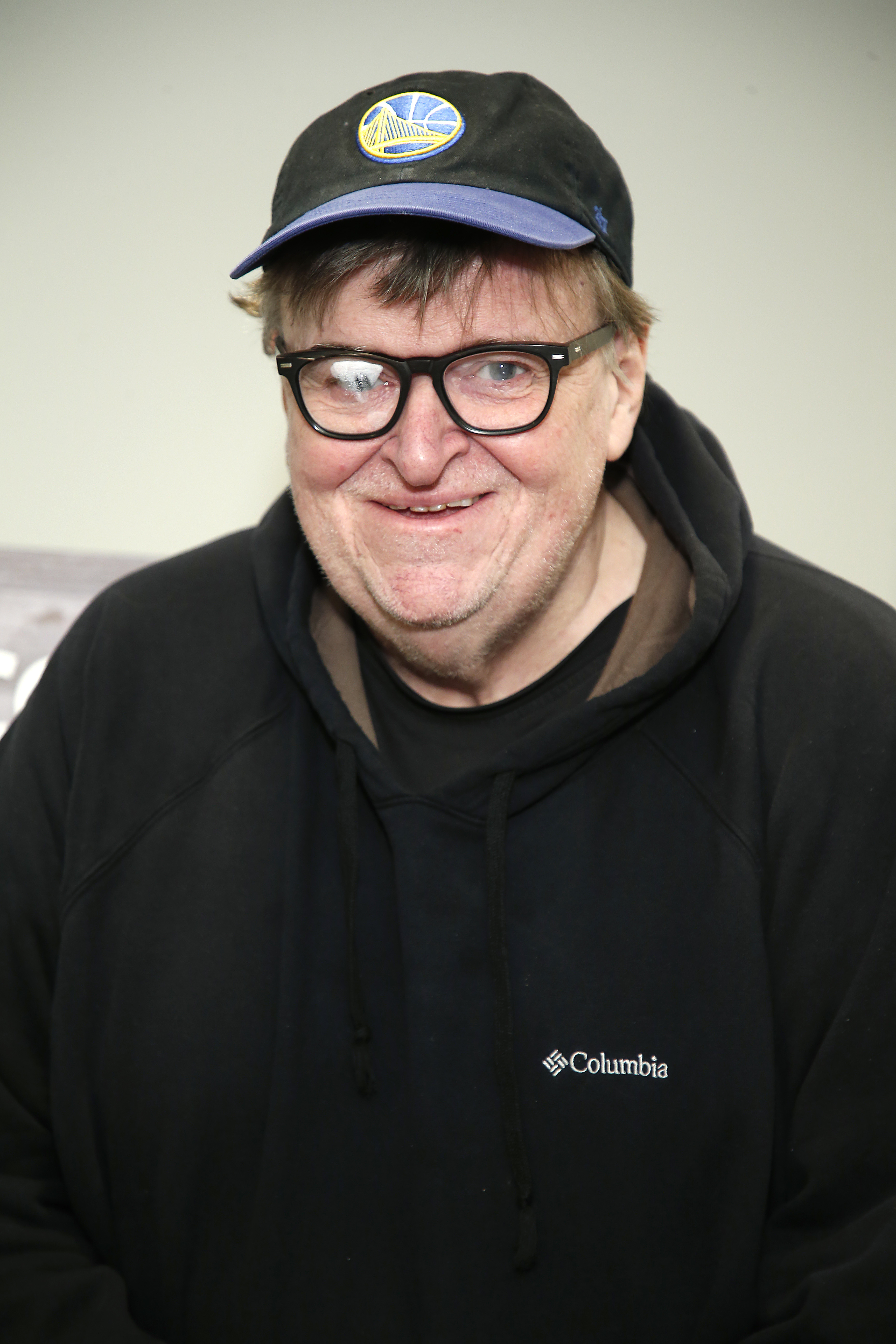 Michael Moore smiling, wearing a baseball cap and glasses, dressed casually in a black hoodie