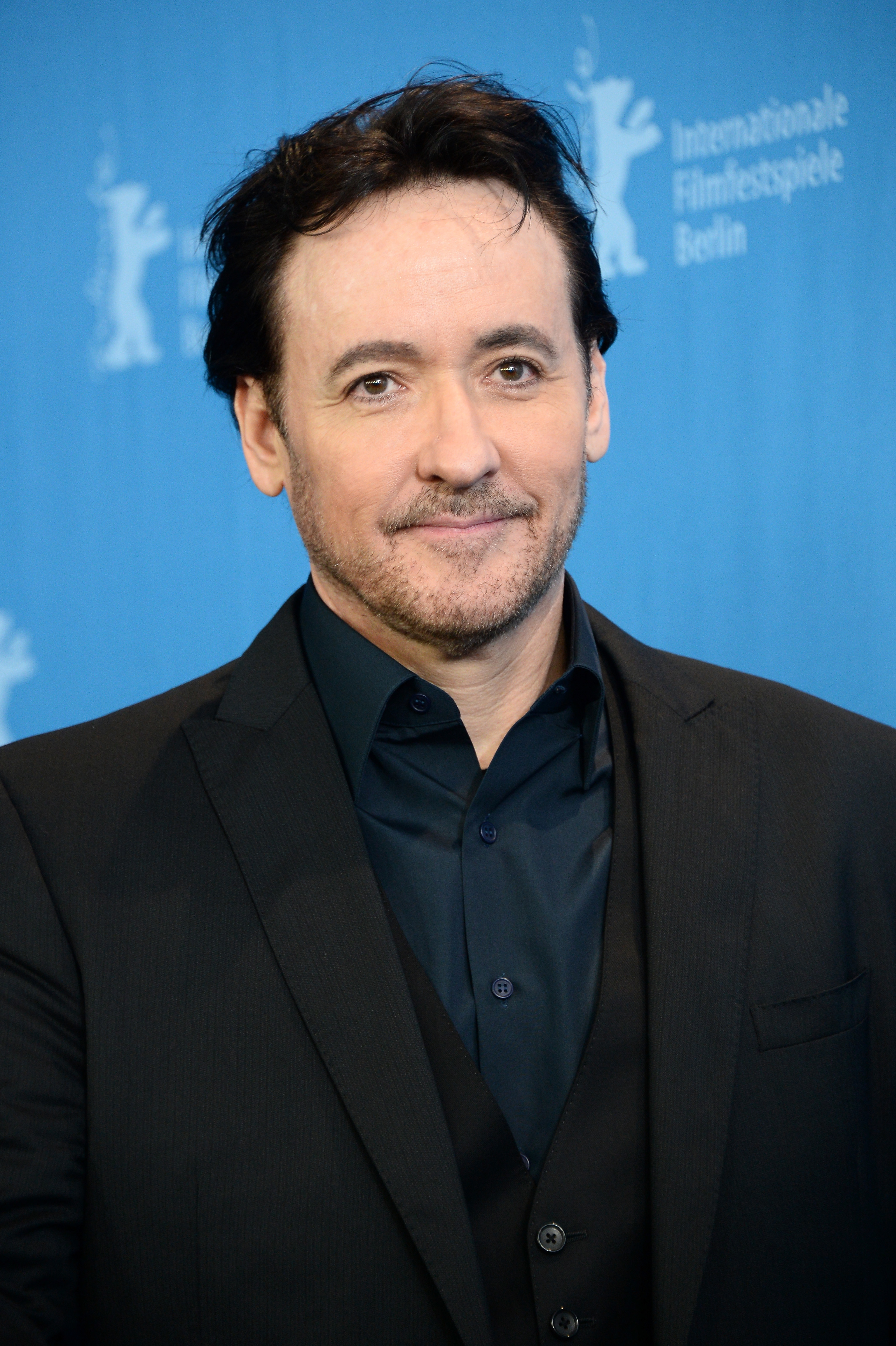 John Cusack in a dark suit and shirt poses in front of a backdrop at the Berlin International Film Festival