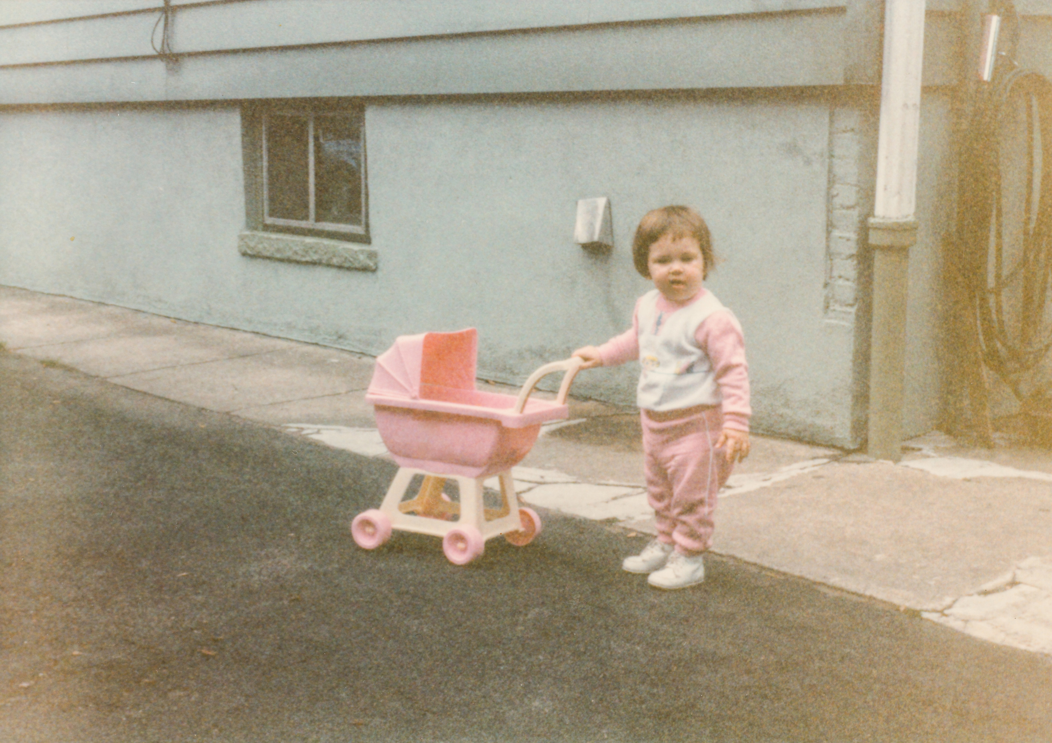 A young child in a tracksuit pushes a toy baby stroller on a paved path next to a building