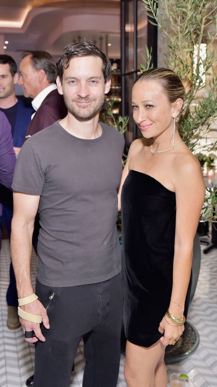 Tobey in a casual t-shirt and pants stands beside Jennifer wearing an elegant, strapless dress at a social event