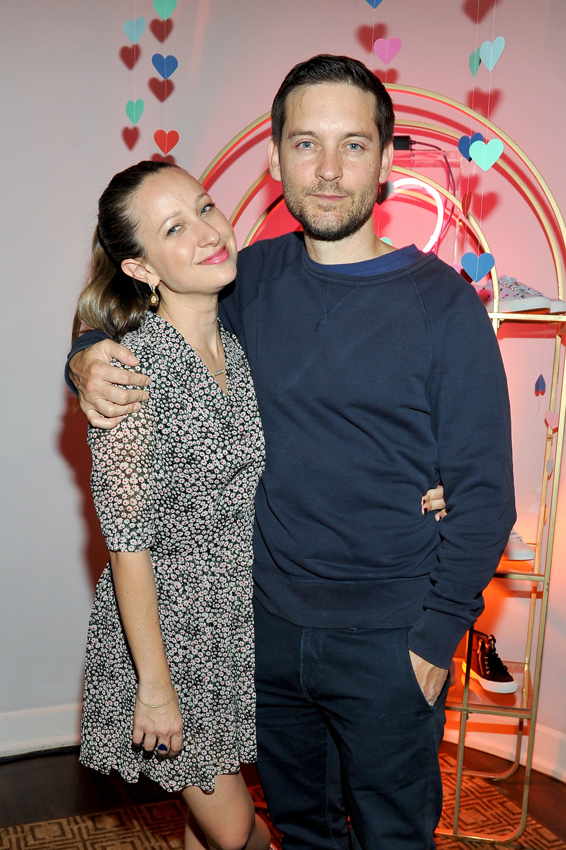 Jennifer Meyer and Tobey Maguire smiling and posing together, with Tobey&#x27;s arm around Jennifer&#x27;s shoulders. They are standing in front of a backdrop with heart decorations