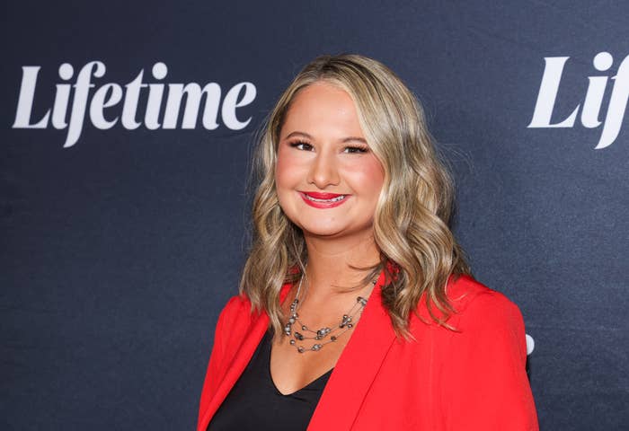 Woman in a black top and red blazer at a Lifetime event, smiling with wavy hair and a layered necklace