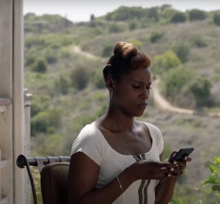 Issa Rae is sitting on a chair outdoors, looking at her phone with a serious expression. She is wearing a white dress. The background features a scenic landscape